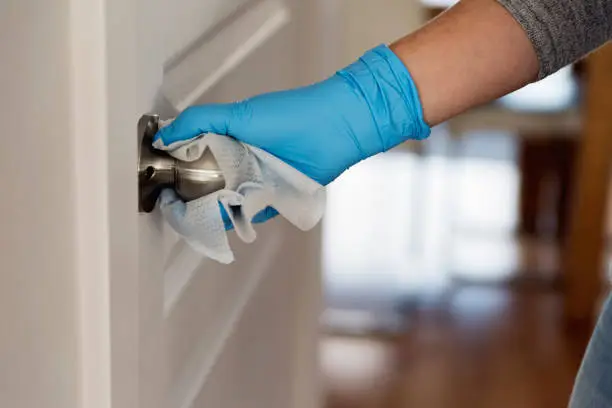 Female hand with blue glove wiping doorknob with disinfectant wipe. Horizontal indoors close-up with copy space.