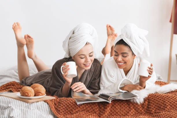 chicas positivas en albornoces y toallas tendidas en la cama - bathrobe women cup towel fotografías e imágenes de stock