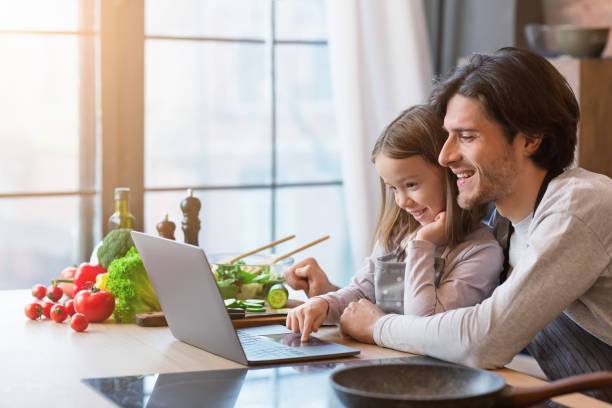 istock Little girl and her dad using laptop at kitchen, looking recipe 1212745960