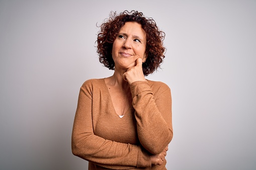 Middle age beautiful curly hair woman wearing casual sweater over isolated white background with hand on chin thinking about question, pensive expression. Smiling with thoughtful face. Doubt concept.