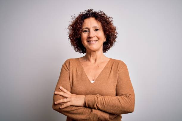 femme de cheveux bouclés de beau âge moyen utilisant le chandail occasionnel au-dessus du visage heureux de fond blanc isolé souriant avec les bras croisés regardant la caméra. personne positive. - senior female photos et images de collection