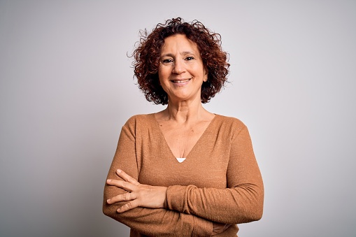 Middle age beautiful curly hair woman wearing casual sweater over isolated white background happy face smiling with crossed arms looking at the camera. Positive person.