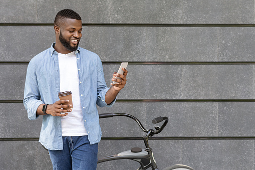Black guy standing next to bicycle, texting on smartphone and drinking takeaway coffee outdoors, copy space