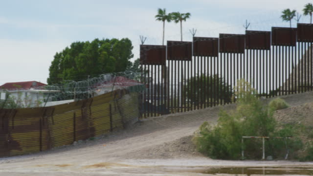 Panning Shot of Parts of the Old and New Steel-Slat Border Wall Topped by Razor Wire (on the US Side) between Mexico and the United States with the Town of Los Algodones and Palm Trees on the Other Side and Bushes in the Foreground