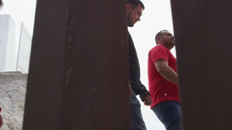 Slow Motion Shot of Five Hispanic Men and a Dog Walking with a Dog on the Mexican Side of the US/Mexican Steel-Slat Border Wall as the Camera Films on the US Side on a Sunny Day