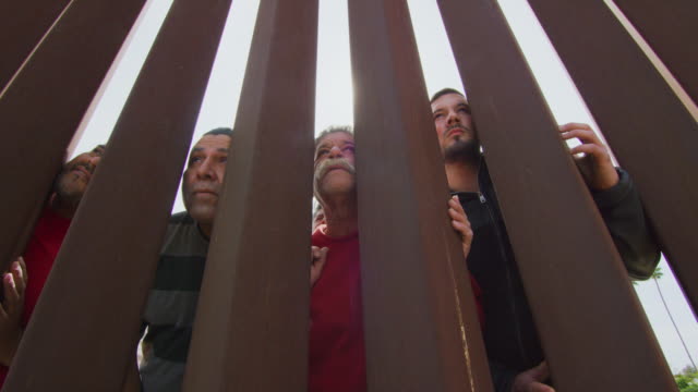 Slow Motion Shot of Four Hispanic Men of Various Ages (on the Mexican Side) Gazing at the United States through the Steel-Slat Border Wall between Mexico and the United States (Shot from the US Side)