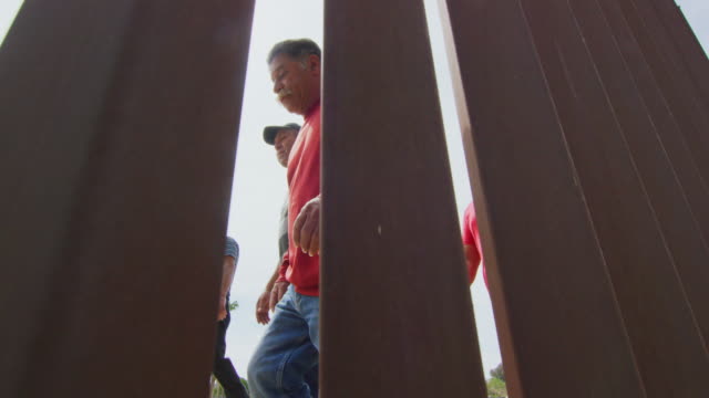 Slow Motion Shot of Five Hispanic Men and a Dog Walking on the Mexican Side of the US/Mexican Steel-Slat Border Wall as the Camera Films on the US Side on a Sunny Day