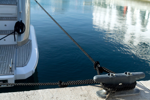 Gray metal mooring post with a black knotted rope in front of the blue water with a moored boat in a sea harbor