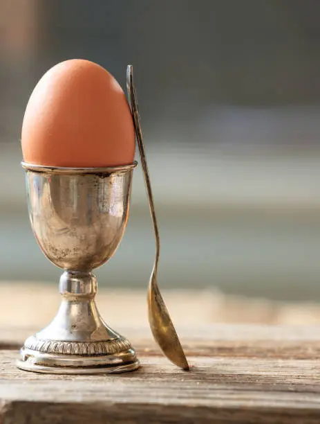Egg in a silver eggcup and a spoon on wooden table. Vertical portrait of the set and a fresh full protein brown egg. Blur background, space.