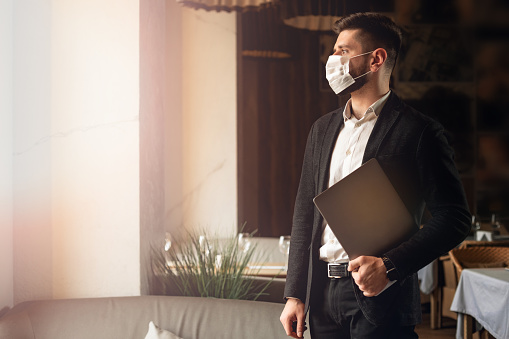 Caucasian adult bearded man indoors in cafe. Lifestyle concept photo with copy space. Picture with handsome guy who using protective face mask. Portrait with gray laptop