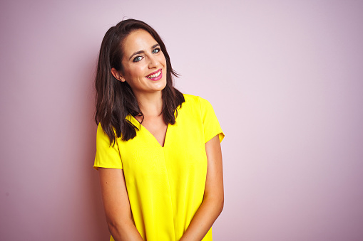 Young beautiful woman wearing yellow t-shirt standing over pink isolated background looking away to side with smile on face, natural expression. Laughing confident.