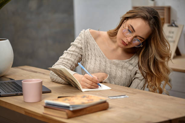istock Lady blogger in glasses, beige sweater. Sitting in kitchen at table, writing in her notebook. Laptop, book, plastic card and pink cup nearby. Close up 1212764241