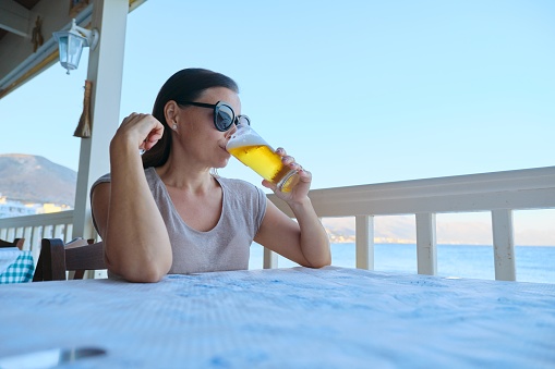Mature beautiful woman resting sitting in sea cafe drinking cold glass of beer. Summer vacation, female enjoys sea sunset landscapes and delicious drink, copy space