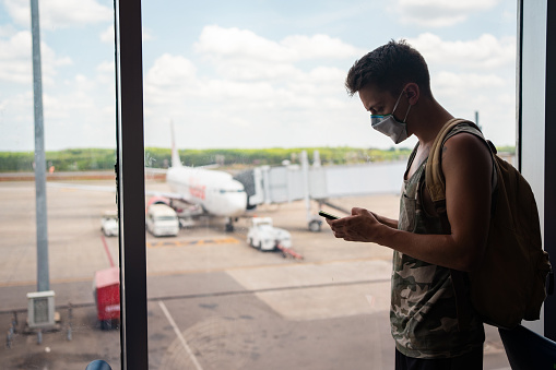 Young man with protective mask is using smart phone while waiting his flight at the airport.