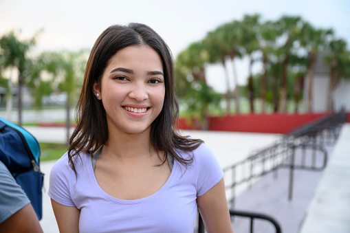 Smiling young Hispanic female teenager with medium-length brown hair pausing to look at camera on her way into school.