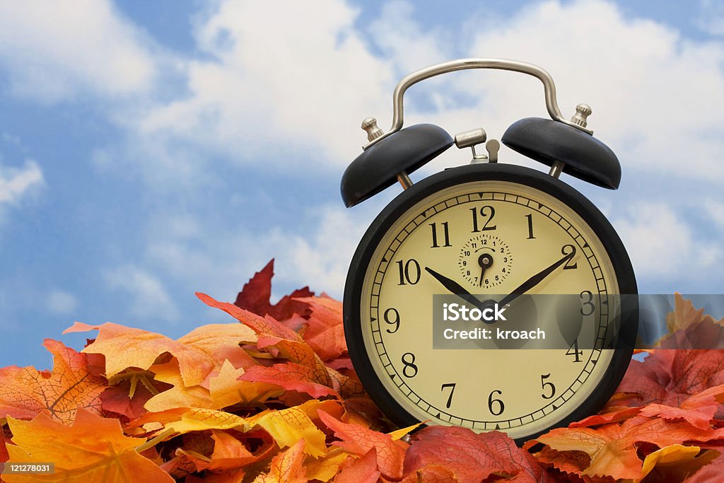 Alarm clock sitting on fall leaves indicating time change Retro clock on fall leaves sky background Aging Process Stock Photo