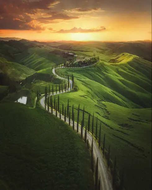 Magical sunset and rural landscape view of Picturesque agrotourism with characterized green top hill farms of olive groves and vineyards typical curved road with cypress at Crete Senesi in Toscana, Italia, Europe