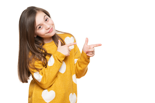 Beautiful young girl in mustard yellow sweater looking at camera, smiling and pointing to the side. Waist up studio shot on white background.