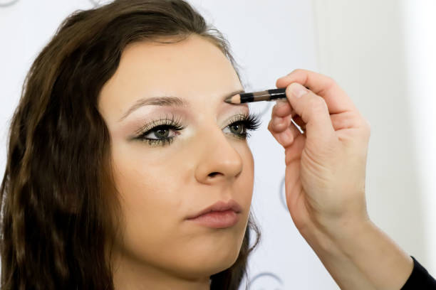 istock Make-up artist applying eye shadow on eyebrow to customer with  eyebrows pen. Profile of beautiful  young woman in beauty studio, close up 1212785725