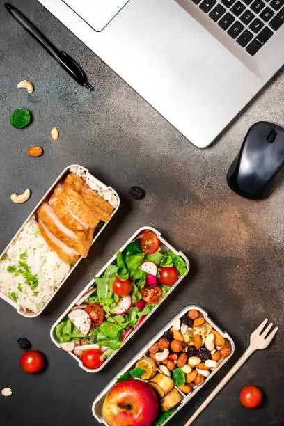 Lunch at the office or workplace. Lunch boxes with chicken, rice, salad, fruits and nuts on a dark desktop, top view. Healthy balanced office food in lunchboxes, flat lay