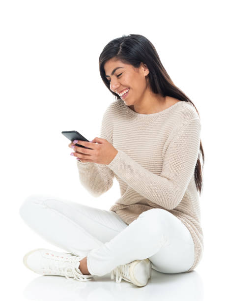 Latin american and hispanic ethnicity young women sitting in front of white background wearing warm clothing and using smart phone Side view of aged 20-29 years old who is beautiful with long hair latin american and hispanic ethnicity young women sitting in front of white background wearing warm clothing who is smiling and using smart phone sitting on floor stock pictures, royalty-free photos & images