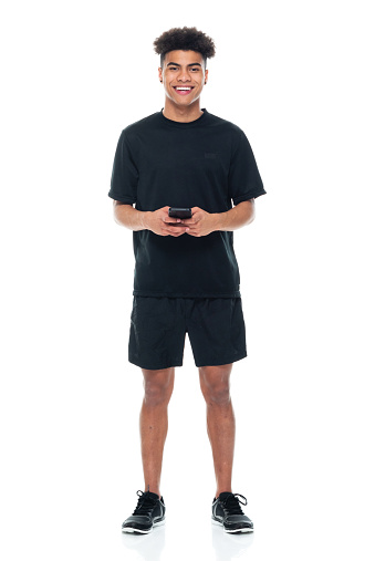 Front view of aged 18-19 years old with curly hair african-american ethnicity male standing in front of white background wearing sports shoe who is smiling and using mobile phone