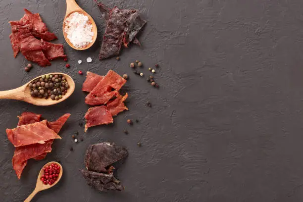 Jerky. Set of various kind of dried spiced meat, diverse peppercorns and salt on wooden spoons  on dark gray background. Top view with copy space. Snack for beer.