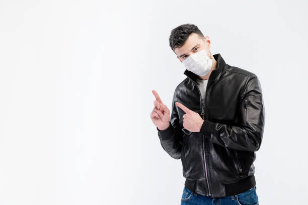 hübscher kaukasischer junger mann in schwarzer jacke und blauer jeans mit einweg-gesichtsmaske. schutz vor viren und infektionen. studioportrait, konzept mit weißem hintergrund. er gestikuliert, kopiert platz für ihr design. - gauze healthcare and medicine disposable bandage stock-fotos und bilder