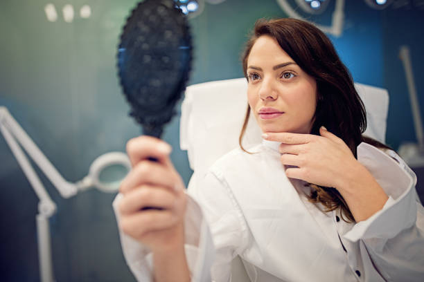 молодая женщина смотрит своим лицом в зеркало после процедуры лечения красоты - plastic surgery botox injection face lift nose job стоковые фото и изображения