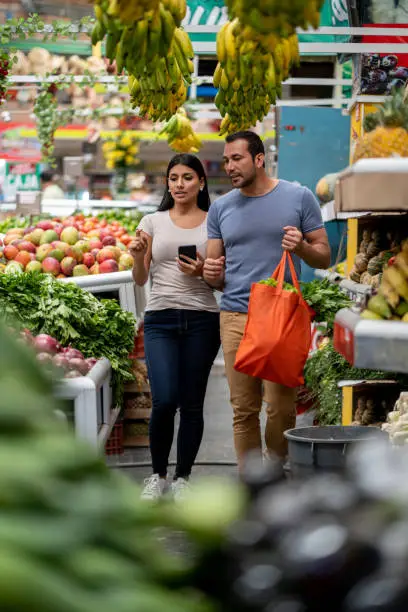 Young couple buying fruits and vegetables while woman looks at shopping list on smartphone's app and man holds a reusable bag
