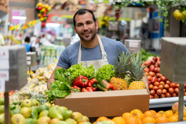 Handsome man working at a farmer's market holding an order for customer in a cardboard box looking at camera smiling Handsome man working at a farmer's market holding an order for customer in a cardboard box looking at camera smiling - Responsible business market vendor stock pictures, royalty-free photos & images