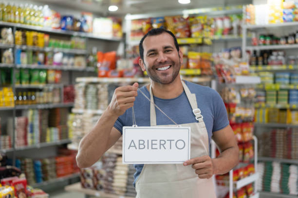 verticale du propriétaire d’entreprise latino-américain d’un petit marché retenant un signe ouvert souriant à l’appareil-photo - commerçant photos et images de collection