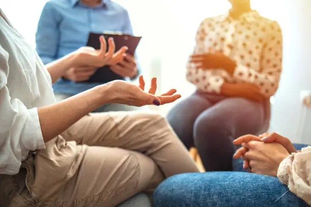 Woman gestures while discussing something difficult during a support group or group therapy session. A mental health professional is taking notes in the background. People at group psychotherapy session indoors