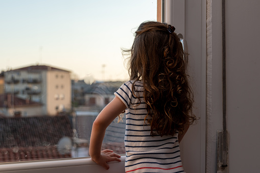 cute little girl spending time looking through the window after a week of quarantine for the coronavirus in italy