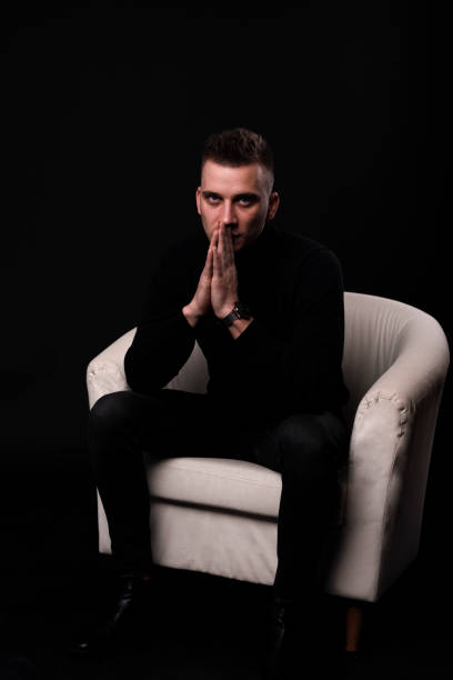 istock dramatic cool guy holding elbow on knee in a fashion pose, sitting on grey background 1212834751