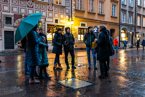Warsaw, Poland - December 22, 2019: Group of tourists people on guided tour with guide at Warszawa old town market square with rain umbrella at night or evening with lights lamps