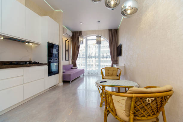 istock Kitchen room with a fresh renovation in beige tones. White kitchen set with kitchen appliances. semicircular window with curtains. TV, sofa and wicker furniture-table and two chairs 1212880698