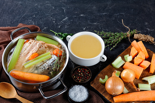 fish stock or soup of salmon, onion, carrot, celery, herbs and spices in a stockpot and in a white bowl on a concrete table with ingredients, free space, horizontal view from above, close-up