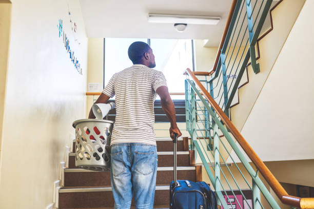 Am I in the right place? Rearview shot of a carefree young man carrying a washing basket and luggage while walking up a flight of stairs inside of an apartment building during the day dorm room photos stock pictures, royalty-free photos & images
