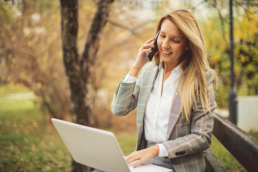 Beautiful young woman taking a break from her job and using laptop and mobile phone in city park for online communication with somebody. She looks very happy and cheerful