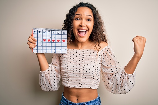 Young beautiful woman with curly hair holding mensturation calendar over white background screaming proud and celebrating victory and success very excited, cheering emotion