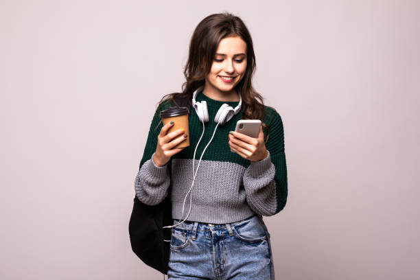 istock Young student woman with backpack and white headset video chatting via mobile phone while standing on a white background. 1212920701
