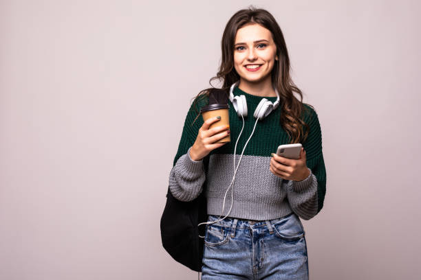 istock Young student woman with backpack and white headset video chatting via mobile phone while standing on a white background. 1212920749