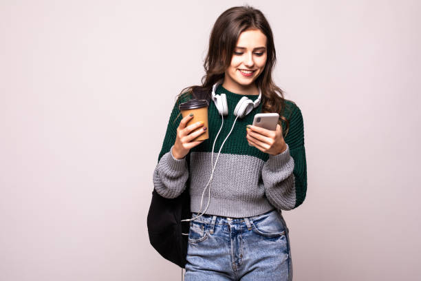 istock Young student woman with backpack and white headset video chatting via mobile phone while standing on a white background. 1212920857