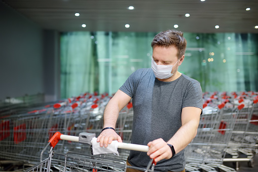 Man wearing disposable medical face mask wipes the shopping cart handle with a disinfecting cloth in supermarket. Safety during coronavirus outbreak. Epidemic of virus covid.