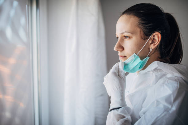 Worried female doctor looking through the hospital window One woman, worried female doctor in protective suit, looking through the hospital window. cleanroom photos stock pictures, royalty-free photos & images