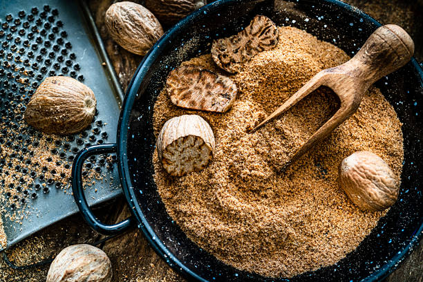 Top view of a rustic wooden table with nutmeg seeds and ground nutmeg. A small grater is beside a container filled with ground and cracked nutmeg seeds. Predominant color is brown. High resolution 42Mp studio digital capture taken with SONY A7rII and Zeiss Batis 40mm F2.0 CF lens