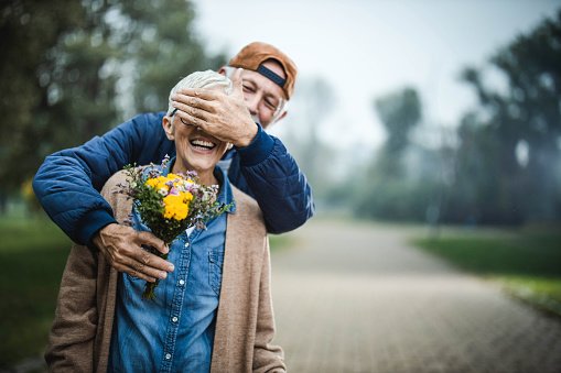 Happy senior man covering his wife's eyes while brining her flower bouquet at the park. Copy space.