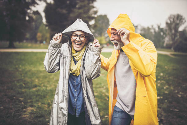 Happy senior couple in raincoats during rainy day in nature. Happy mature couple covering their heads with hood during rainy day in the park. raincoat stock pictures, royalty-free photos & images