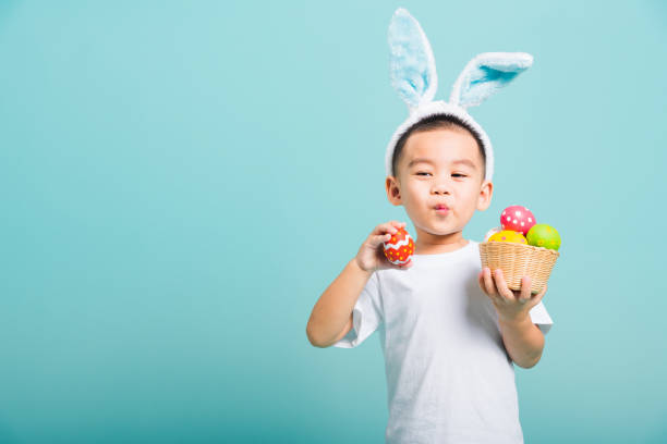 azjatycki cute little boy dziecko uśmiech rozpromieniony sobie uszy króliczka i biały t-shirt, stojący trzymać kosz z pełnymi pisankami. a druga ręka trzyma jajko wielkanocne - easter baby rabbit eggs zdjęcia i obrazy z banku zdjęć